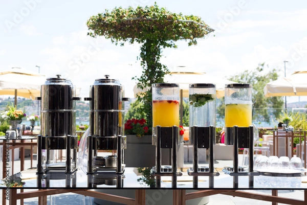 Fototapeta An elegant outdoor refreshment setup featuring glass dispensers filled with fruit-infused water and juices, perfect for a sunny garden gathering or catered event.