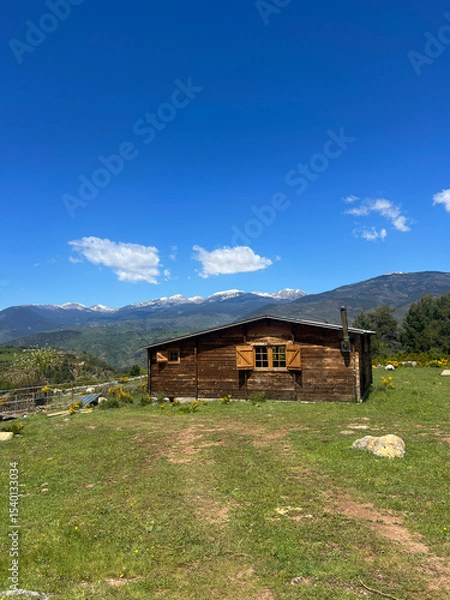 Fototapeta A wooden cabin with Snow-Capped Peaks in the mountains on a sunny day. Rustic Cabin. Scenic view of rolling hills, mountains, and a cloudy sky under sunlight.
