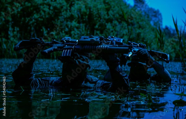 Obraz Two armed soldiers in camouflage, carrying rifles above water, wade slowly through a dense, swamp-like area surrounded by greenery.