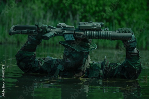 Obraz Camouflaged soldier with face paint held rifle above head while wading through water in a dense forest environment under daylight.