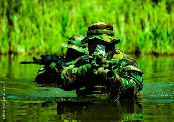 Obraz Soldiers in camouflage uniforms with rifles navigate through water in a dense green vegetation setting, demonstrating stealth and tactical movement.