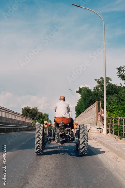 Fototapeta A farmer from behind slowly drives an old tractor across a bridge, typical of a late summer afternoon, and conveys a sense of homecoming after a long day of work in the fields.