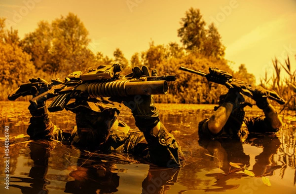 Obraz Two camouflaged soldiers wade through water holding rifles above their heads, with trees in the background on a clear day.