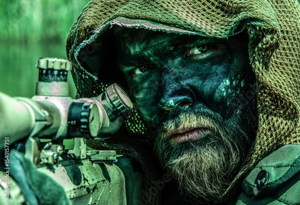Obraz A sniper in a camouflage netting hood with face paint peers through a rifle scope in a dense green environment.
