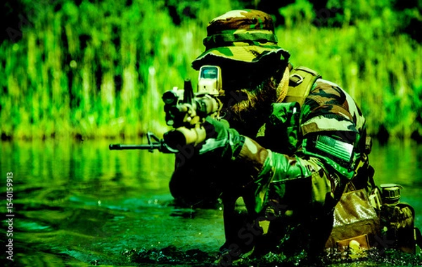 Obraz Soldier in camouflage uniform and hat aiming a rifle with a scope while wading through a green forested water area.