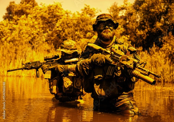 Obraz Two armed soldiers in camouflage and tactical gear wade through water, holding rifles, with trees and foliage in the background.