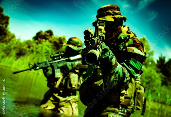 Obraz Two soldiers in camouflage uniforms and helmets aim rifles in a lush, green landscape, partially submerged in water with trees in the background.