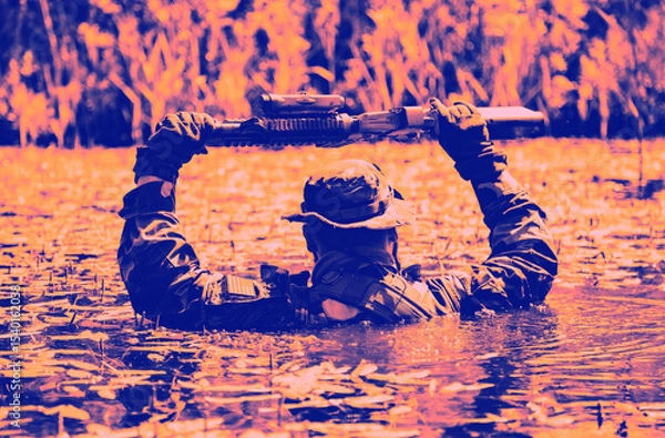 Obraz A soldier wearing camouflage holds a firearm above his head while wading through water surrounded by reeds in a dramatic color filter.
