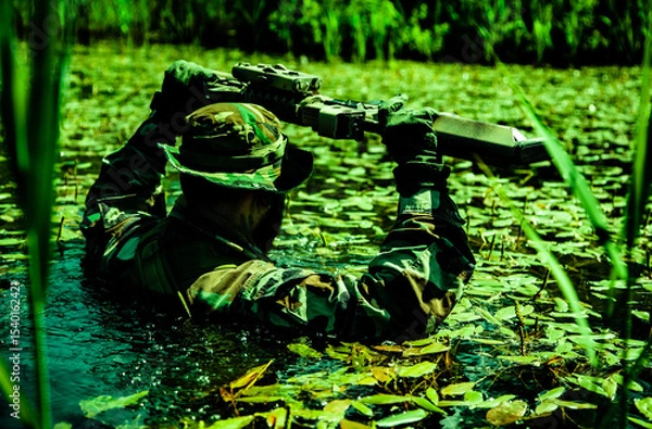Obraz Soldier in camouflage wades through a swamp holding a rifle above the water surface surrounded by aquatic plants and greenery.