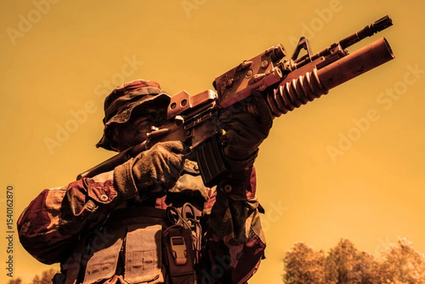 Obraz Soldier in camouflage attire and field helmet aiming a rifle with mounted optic under a yellow-orange sky, with trees in the background.