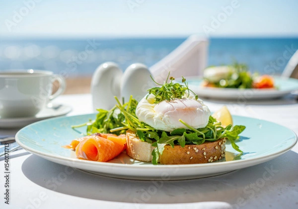 Obraz vibrant breakfast plate featuring poached egg, smoked salmon, and arugula on toasted bread against a seaside backdrop with sunlight reflecting off the ocean