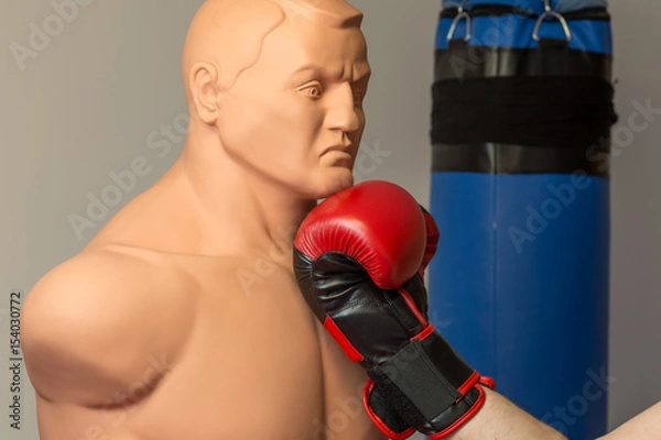 Fototapeta Red boxing gloves punching an uppercut on the chin of a dummy mannequin doll on a gym with a blue punching bag on the background