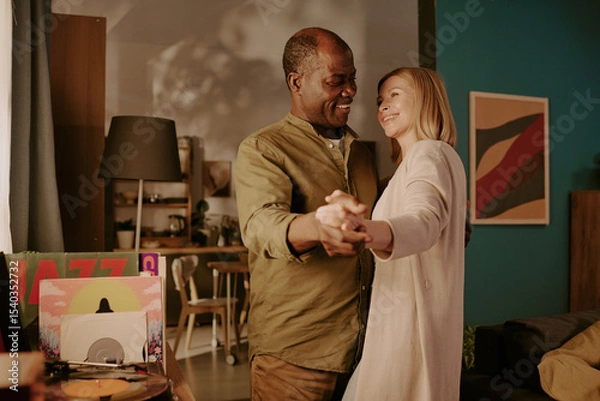 Obraz Senior Black man and senior Caucasian woman dancing together in living room, smiling and holding hands, enjoying romantic moment, standing close and looking at each other