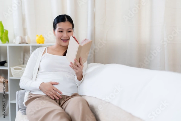 Fototapeta Asian pregnant woman relaxing on sofa smiling holding book reading calmly with hand resting on belly wearing comfortable brown pants and white sweater showing peaceful happiness before childbirth