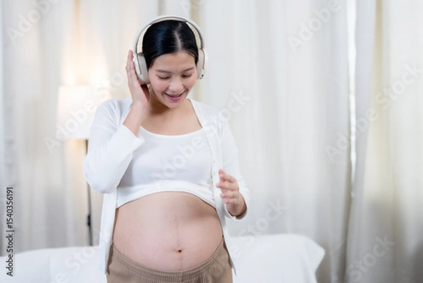 Fototapeta Asian pregnant woman smiling wearing headphones standing happily enjoying music with hand resting on belly expressing excitement bonding emotional connection with unborn baby at cozy home