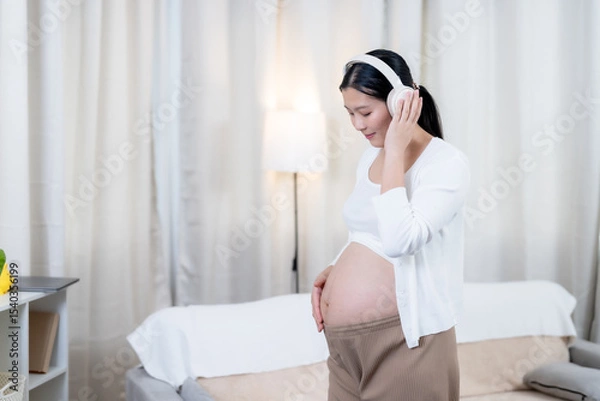 Fototapeta Asian pregnant woman standing wearing headphones smiling touching belly enjoying music connecting emotionally with unborn baby in cozy living room wearing white sweater and brown pants