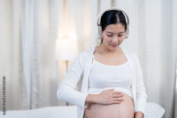 Fototapeta Asian pregnant woman wearing headphones standing calmly touching belly eyes closed enjoying peaceful music creating emotional connection with unborn baby preparing for happy motherhood journey ahead