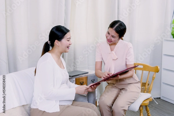 Fototapeta Asian pregnant woman smiling warmly while discussing newborn insurance with female agent reviewing documents showing friendly interaction support and shared understanding in cozy home setting