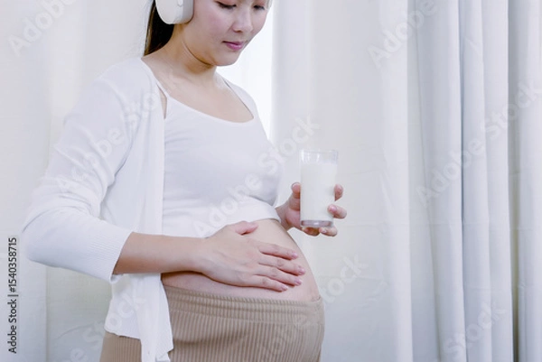 Fototapeta Asian pregnant woman standing peacefully holding glass of milk with one hand resting on belly wearing headphones showing healthy routine and emotional bonding with unborn baby in quiet home