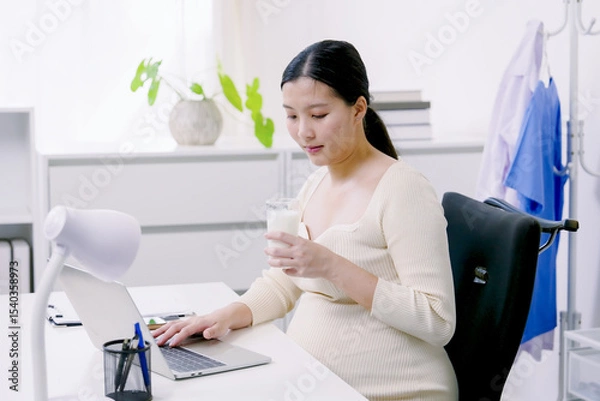 Fototapeta Asian pregnant woman working at desk using laptop holding glass of milk in home office wearing cream knit top showing balanced lifestyle between career and health during late pregnancy