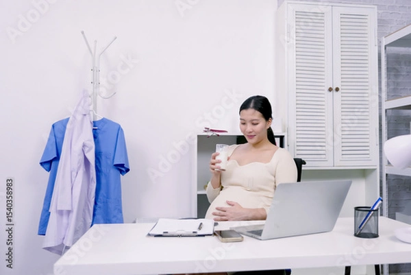 Fototapeta Asian pregnant woman taking short break at office sipping milk while resting hand on belly showing care for health and baby while maintaining balance during intense workday in final pregnancy stage