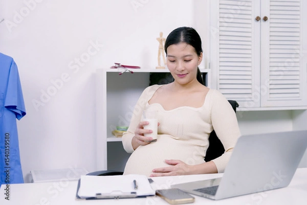 Fototapeta Asian pregnant woman relaxing with glass of milk in office environment resting hand on belly showing self-care nurturing unborn baby during late pregnancy while staying responsible at workplace