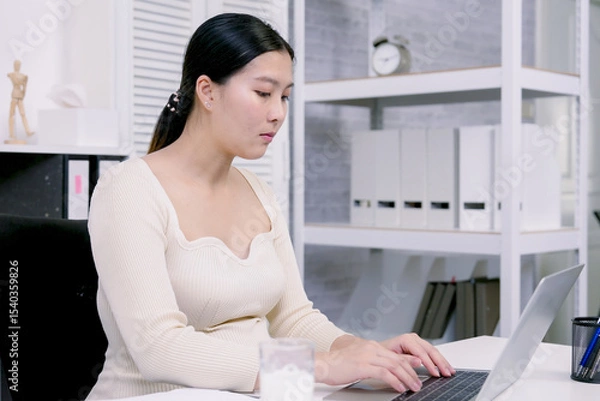Fototapeta Asian pregnant woman working at office desk focused on laptop with glass of milk nearby showing professional discipline and balance between career and personal health close to delivery time