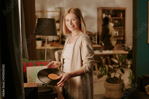 Obraz Caucasian senior woman smiling while holding vinyl record near turntable, standing in cozy living room with another senior in background, engaging in relaxed home activity