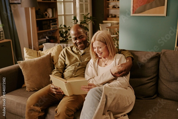 Obraz Senior Black man and senior Caucasian woman sitting on sofa smiling and reading book together, man embracing woman, couple enjoying romantic moment in cozy living room setting