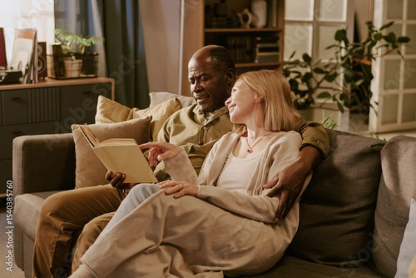 Obraz Senior Black man and senior Caucasian woman sitting together on sofa reading book, smiling and embracing, showing romantic connection and enjoying shared moment in cozy living room