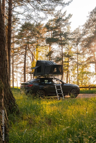 Obraz A modern SUV with a foldable rooftop tent parked in a forest. Tall pine trees surround the vehicle, with sunlight filtering through the branches.