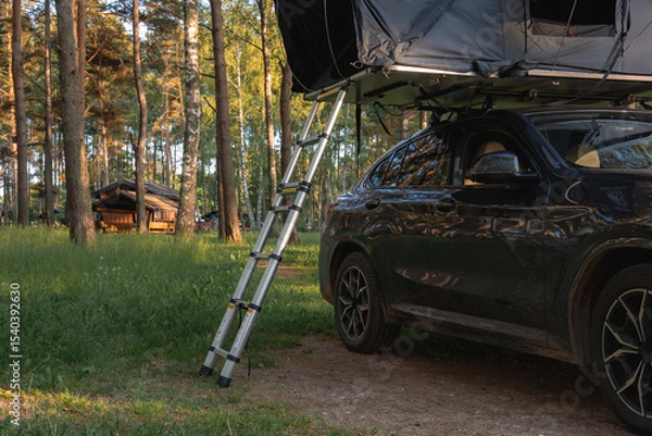 Obraz A modern SUV with a rooftop tent and ladder is parked in a forest of tall pine trees. Sunlight filters through, with a wooden cabin in the background.