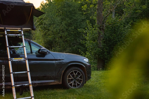Obraz A dark colored SUV with a foldable rooftop tent and ladder parked on grass in a forest. Dirt on the vehicle suggests recent off road travel.