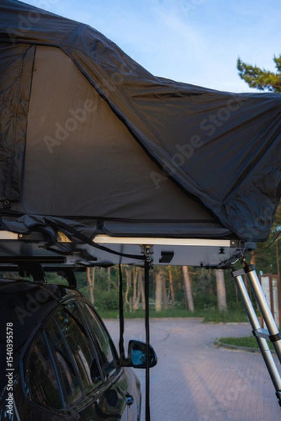 Obraz A modern SUV with a fully extended rooftop tent parked on a paved surface in a forest. Natural light illuminates the trees and camping setup.