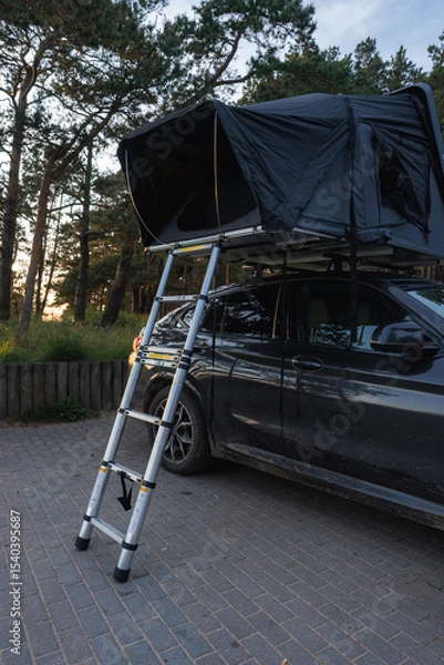 Obraz A modern SUV parked on a paved surface in a forest, featuring a black rooftop tent with a telescopic ladder. Tall trees and sunlight create a serene setting.