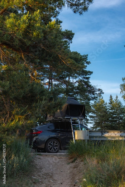 Obraz A modern SUV with an open foldable rooftop tent is parked among tall pine trees. A metal guardrail is visible, suggesting a nearby road or path.