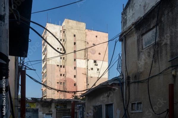 Fototapeta Petah Tikva, Israel, 12  JUNE 2025,  A Tall, Dilapidated Building With Exposed Wires And Smaller Buildings In The Foreground.