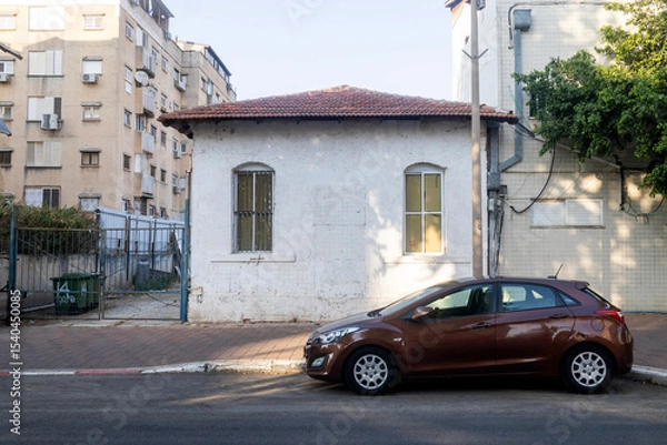 Fototapeta Petah Tikva, Israel, 12  JUNE 2025,  A Small old White House With A Red Tiled Roof Sits Next To A Parked Brown Car. Between last and future