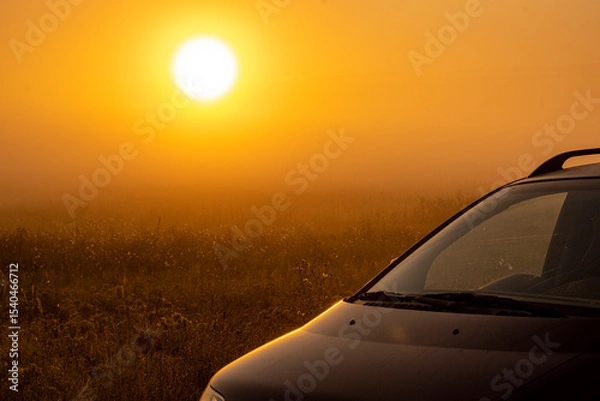 Fototapeta A fascinating panorama of a field unfolding under the morning sky at sunrise. A car is parked on an autumn meadow, bathed in the first rays of light.