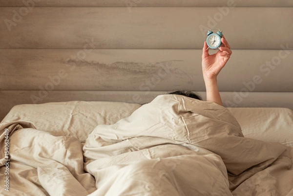 Obraz A hand holding an alarm clock emerging from a cozy bed.