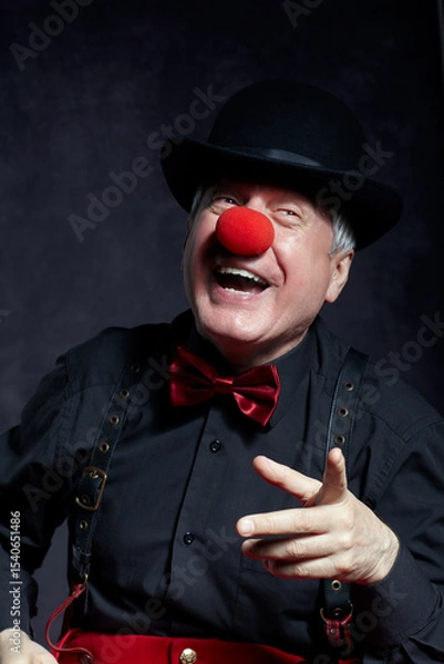 Fototapeta Portrait of a clown on dark background. An elderly man with red clown nose and bowler hat on head. Dressed in red pants and a black shirt with suspenders. The clown points to the side.