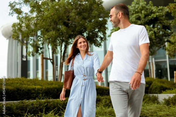 Obraz Couple enjoys a leisurely stroll through urban greenery during a bright afternoon in a modern city setting
