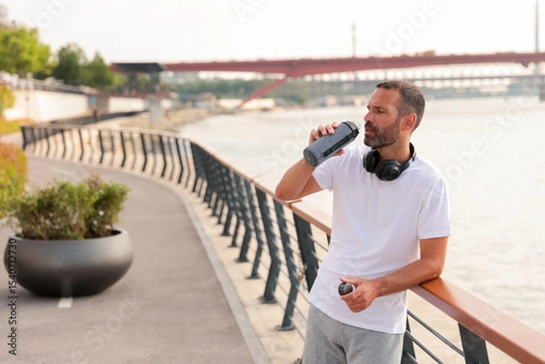 Obraz Man enjoying water break while jogging near a river in a sunny afternoon by the waterfront promenade