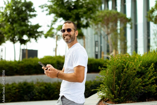 Obraz Smiling man in sunglasses enjoys leisurely stroll while texting in sunny urban park setting