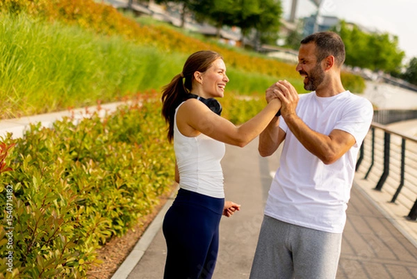 Obraz Couple enjoying a playful moment while jogging along a scenic urban waterfront pathway in bright morning light