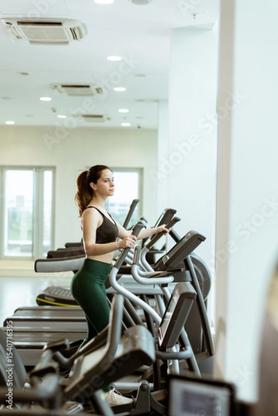 Obraz Woman exercising on elliptical in modern fitness center with vibrant natural light entering through large windows