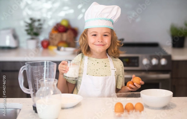 Fototapeta Funny kid with eggs at kitchen. Chef kid boy baking on the kitchen. Child chef cook prepares food at kitchen. Kids cooking. Teen boy with apron and chef hat preparing a healthy meal.