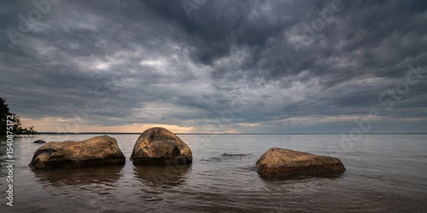 Obraz Evening lake landscape. Large boulders jut out of calm water near the shore of a large reservoir under a dramatic cloudy sky. Widescreen side view in 10x5 format