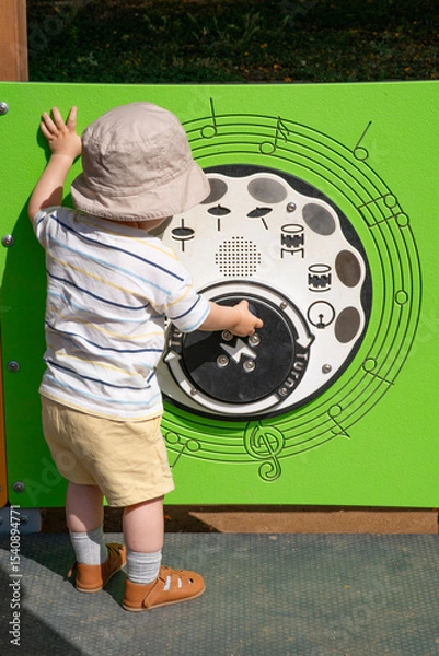 Obraz Musical activity panel at an outdoor playground. The child is interacting with bright green sound panel featuring musical notes and drums icons. Scene captures a moment of early sensory exploration. 
