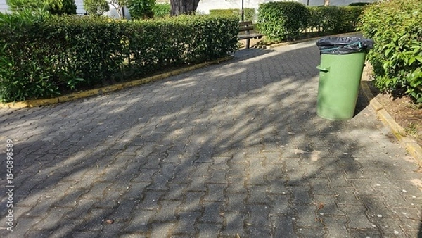 Fototapeta Park pathway with green trash bin and wooden benches surrounded by hedges